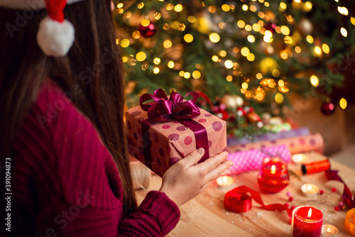 Close-up view of a beautiful gift box in hands of young girl in burgundy sweater and christmas hat, sitting near the table, full of decoration. Cozy atmosphere, bright warm lights.