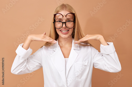 People and lifestyle concept. Isolated shot of funny cheerful nerdy young Caucasian woman ophthalmologist wearing white lab coat and two pairs of eyeglasses, showing teeth braces, smiling broadly