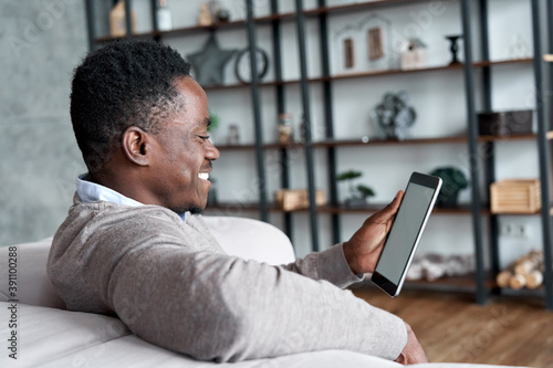Happy smiling african american man holding digital tablet computer sitting on couch at home. Young adult black guy using apps on tab technology device watching videos or browsing internet.