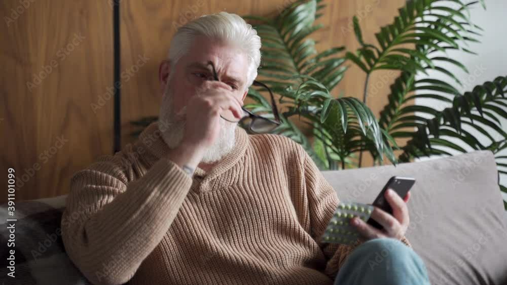 An elderly man is sitting alone on the couch a patient is wearing glasses reading instructions on a smartphone how to take pills An elderly man is sitting alone on the couch a patient is wearing glasses reading instructions on a smartphone how to take pills
