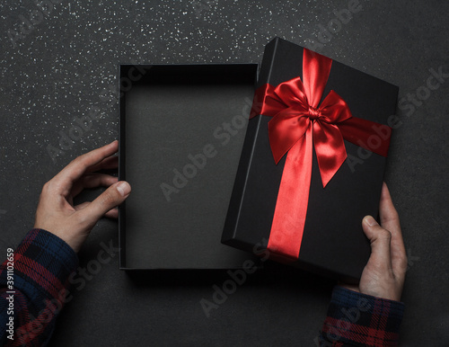 Hands in a shirt open a black gift box with a red bow on a black granite background..