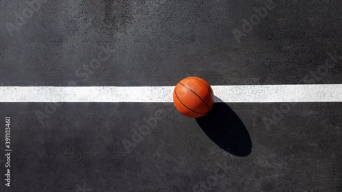 orange striped basketball ball stands on the white line marking the playing field with asphalt tarmac, sports object lit by sun light on playground top view with copy space, nobody.