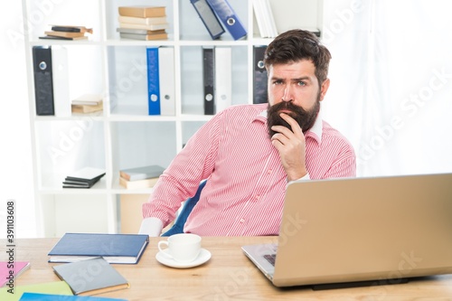 Bearded man with serious look sit at managers desktop in modern work room, office