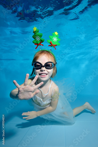 Portrait of a little girl with Christmas decorations in the form of a Christmas tree on her head. She poses for the camera under the water in the pool on a blue background and smiles. Vertical view