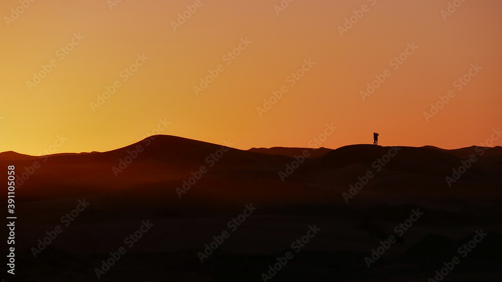 Silhouettes of tourist couple enjoying the colorful sunrise in the sand dunes of Erg Chebbi near Merzouga in southern Morocco, Africa.