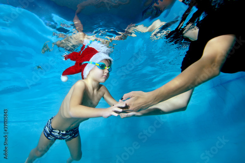 A little boy in a Santa Claus hat swims under the water and reaches for his mother's hands lowered into the water. The concept of love and care. Portrait. Horizontal orientation