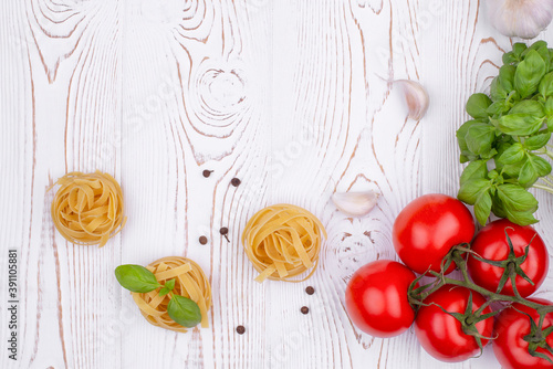 Top view raw tagliatelle pasta with fresh basil, garlic and tomatoes on a rustic white table, flat lay, copy space.