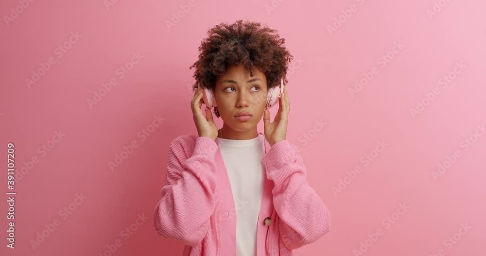 Serious satisfied young Afro American woman enjoys every tune of music wears stereo headphones dressed in casual clothing catches each bit of melody poses against pink background. Hobby concept Serious satisfied young Afro American woman enjoys every tune of music wears stereo headphones dressed in casual clothing catches each bit of melody poses against pink background. Hobby concept