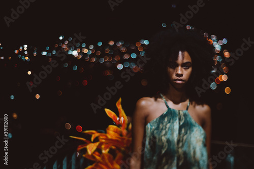 A shot in a dark key of a young ravishing Brazilian female in a simple sundress standing on her house balcony at warm night, with a cityscape behind her, totally blurred into bokeh bulbs