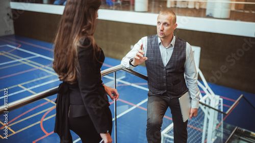 Fototapeta Naklejka Na Ścianę i Meble -  A handsome mature man entrepreneur with a closed laptop in his hand is having a business conversation with a businesswoman indoors of modern office recreation area with a sports field behind them