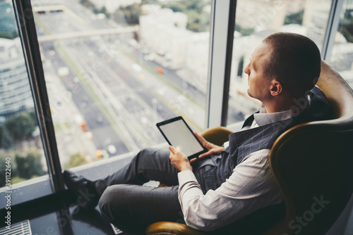 Side view of a pensive mature man entrepreneur in a plaid vest, sitting near the window on an orange armchair in an office cabinet on a top floor of a business high-rise with a digital tablet in hands