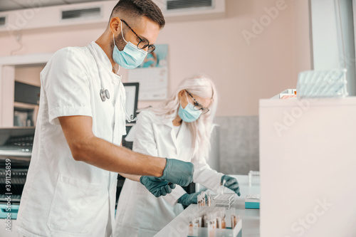 Two focused scientists with rubber gloves and face masks holding test tubes with blood and doing research for cure for corona virus.