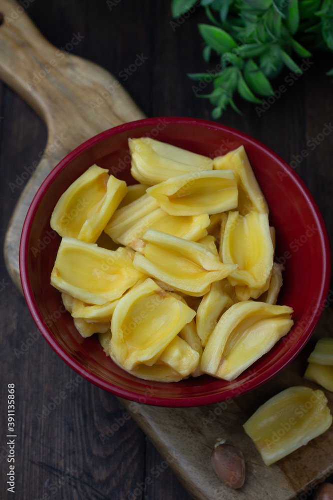 Buah Nangka or Jackfruit, one of tropical fruits popular in Indonesia. Cut into half on red bowl and the seeds at behind. Dark wooden background. Buah Nangka or Jackfruit, one of tropical fruits popular in Indonesia. Cut into half on red bowl and the seeds at behind. Dark wooden background.