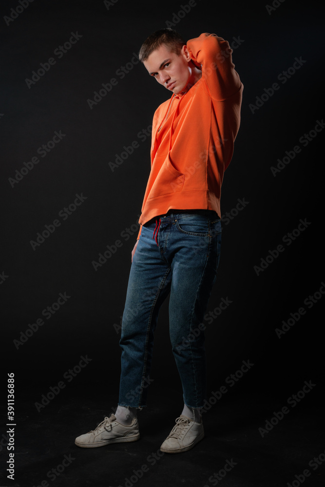 Young handsome man in jeans and orange hoodie posing while standing on a black background. Young handsome man in jeans and orange hoodie posing while standing on a black background.