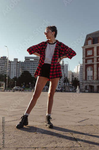 Young beautiful brunette woman in casual plaid red shirt standing, black backpack, outdoors in the city, fashion concept, happy woman, copy space, social media, urban style