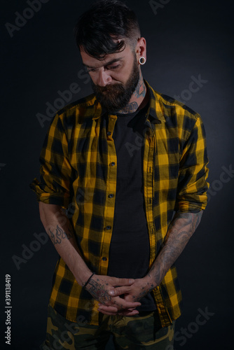 Attractive man with tattoos posing dressed in yellow and black checkered shirt on a black background © qunica.com