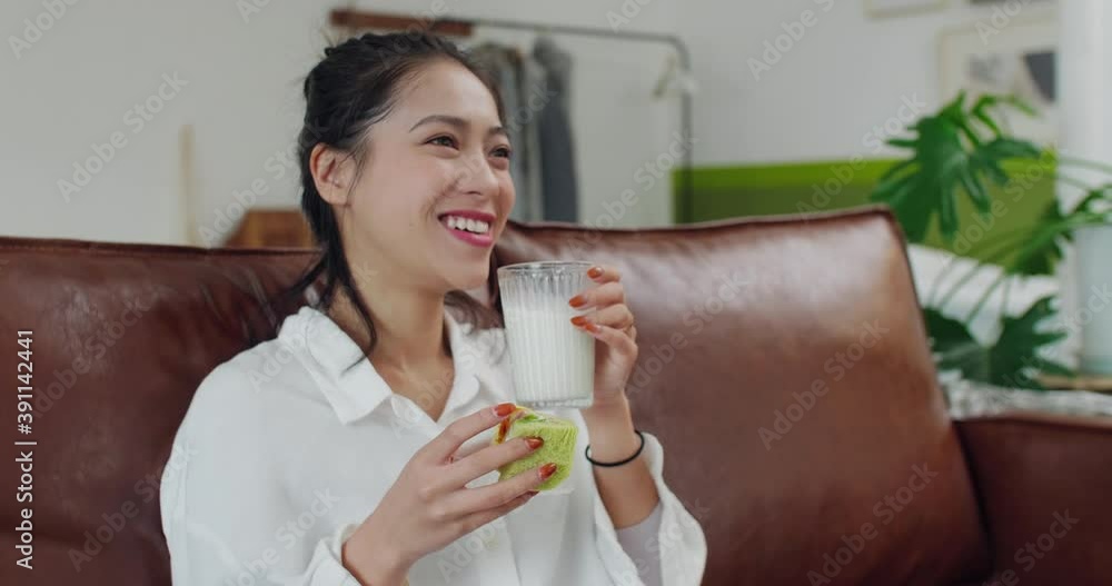 Side view of happy young asian woman sitting in sofa having breakfast watching TV laugh relax lifestyles at home 4k footage Side view of happy young asian woman sitting in sofa having breakfast watching TV laugh relax lifestyles at home 4k footage