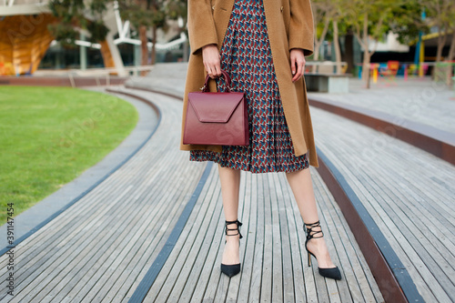 Young fashionable woman wearing beige wool coat, pleated midi skirt and black high heel shoes. She is holding leather handbag in hand. Street style. 