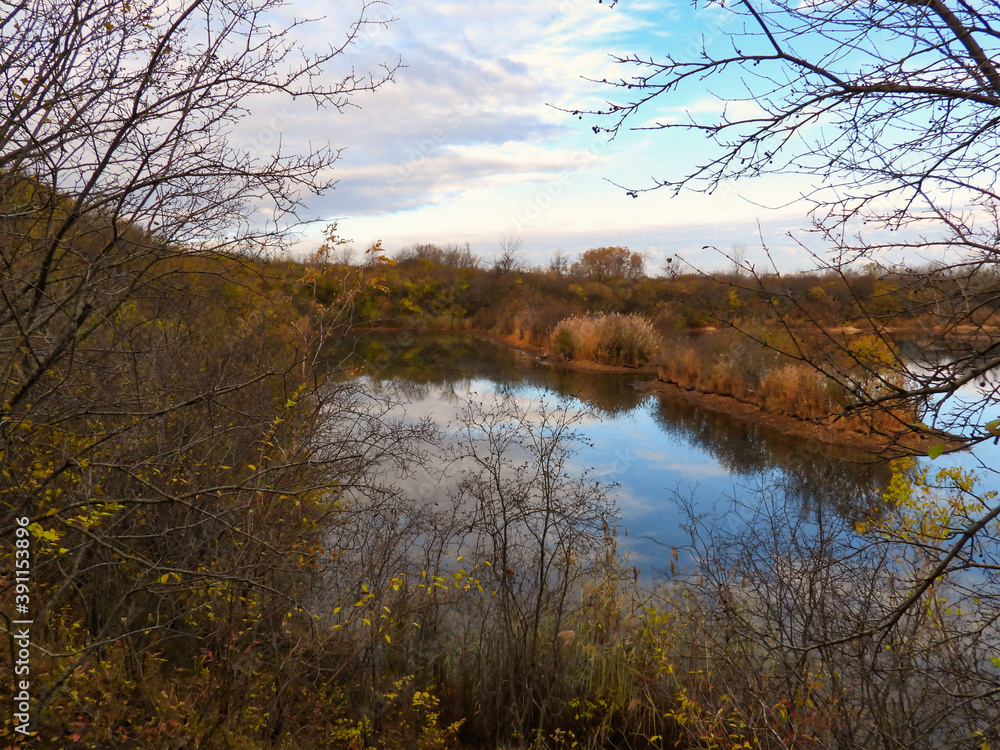 Looking through bare tree branches on a early November, late autumn day to the lake below reflecting the blue sky and clouds with foliage in fall colors on the shore Looking through bare tree branches on a early November, late autumn day to the lake below reflecting the blue sky and clouds with foliage in fall colors on the shore