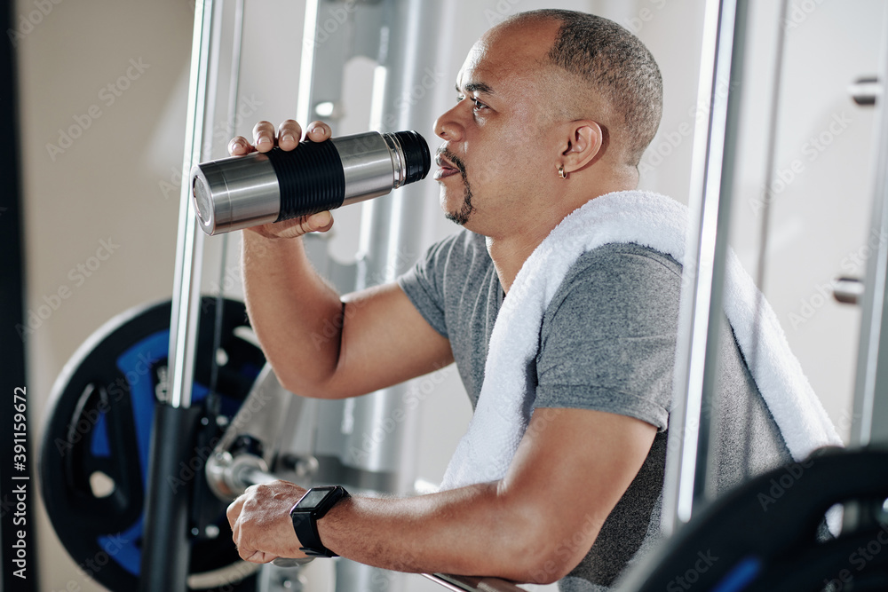 Mature man resting between sets, leaning on barbell and drinking vitamin water from thermos Mature man resting between sets, leaning on barbell and drinking vitamin water from thermos