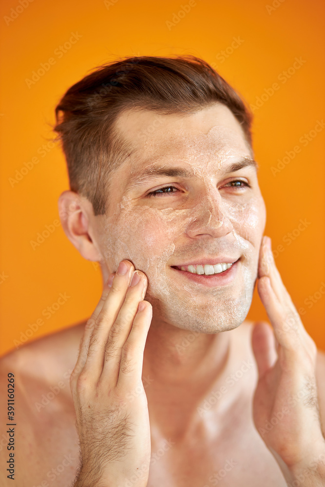 Obraz premium good-looking caucasian man cares for his skin, moisturizes it with professional cosmetics product, isolated orange background