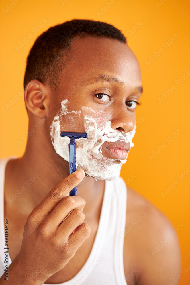 young afro american on yellow background in foam shaving with a razor, portrait of handsome guy posing at camera