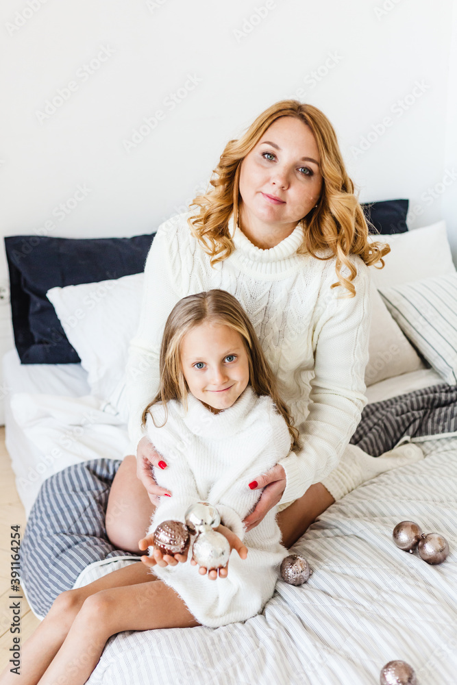 Mom and daughter blonde hair and white sweaters lie on the bed, light up, say toys for Christmas and new year tree, Christmas tree decoration Mom and daughter blonde hair and white sweaters lie on the bed, light up, say toys for Christmas and new year tree, Christmas tree decoration