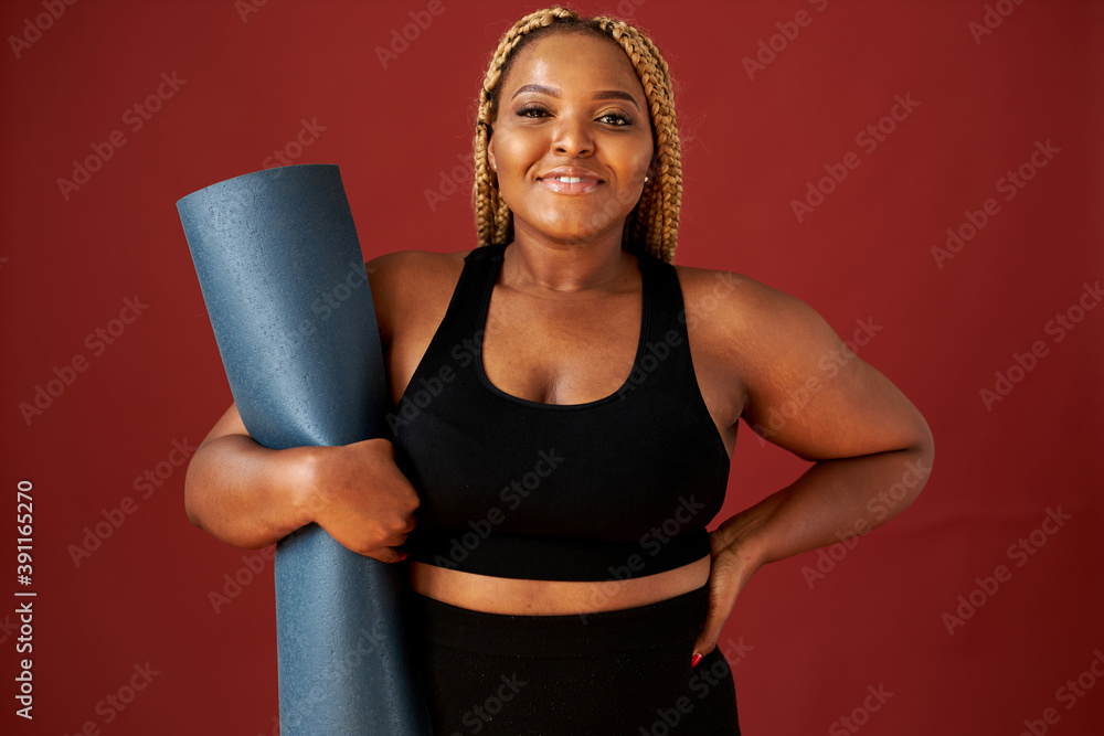 portrait of beautiful chubby african woman going to do sport exercises alone, hold blue mat in hands, posing at camera, wearing black top and leggins - obrazy, fototapety, plakaty 