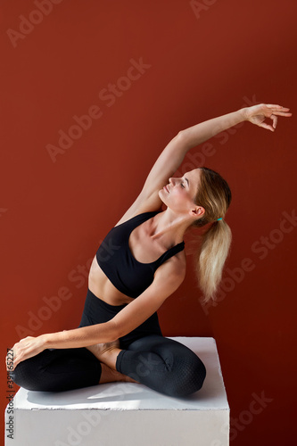 flexible fit woman is stretching while sitting on box isolated on red background, slender female in black sportswear lead healthy lifestyle