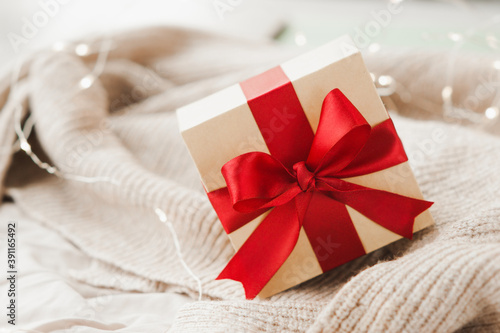 stylish square beige box with red festive satin ribbon on beige crumpled blanket and sparkling garland on background, selective focus © Aleksandra