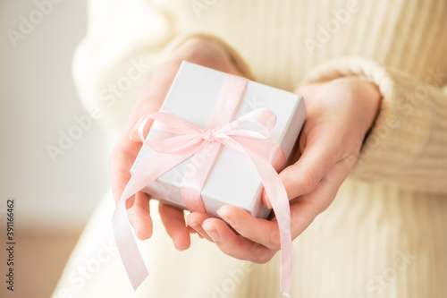 A woman in a sweater gives a gift in a white holiday packaging box with a pink ribbon. Winter festive cozy life style. Romantic present in female hands for christmas, new year