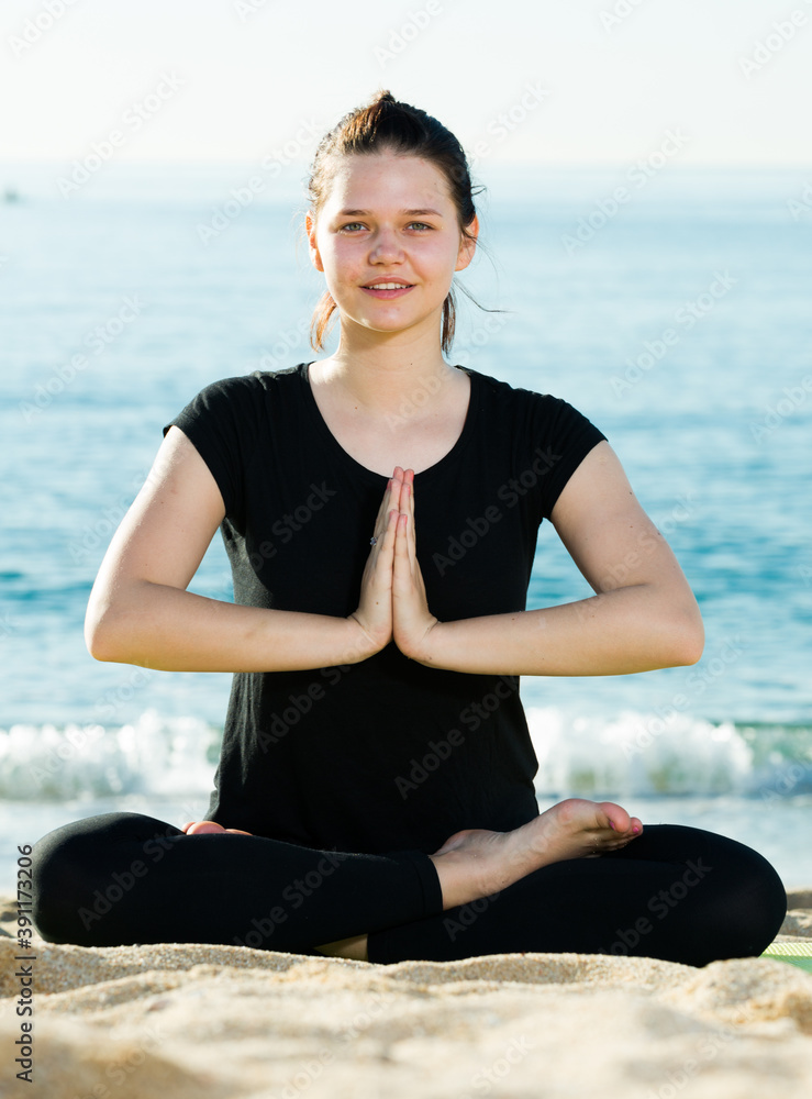 Fitness woman in black T-shirt is sitting and doing yoga on the beach.