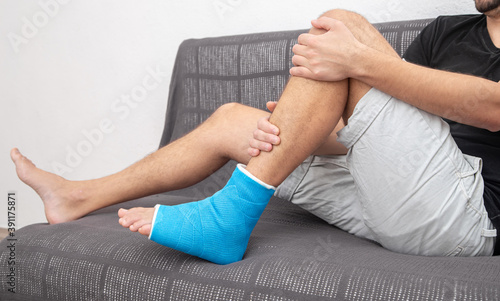 Man with broken leg in blue splint for treatment of injuries from ankle sprain on couch at home. © puhimec