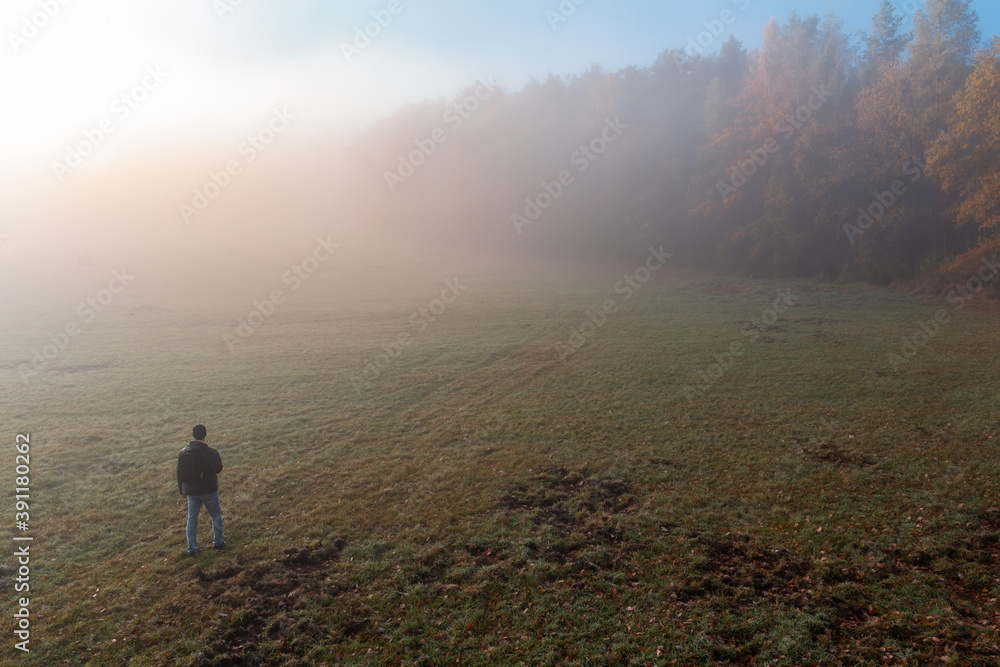 Aerial view young man walking on meadow with autumn forest in freeze misty morning fog. Czech landscape