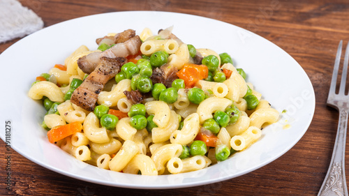 Traditional italian chifferini pasta with peas, carrots and toasted pancetta on a plate on a rustic wooden table, background