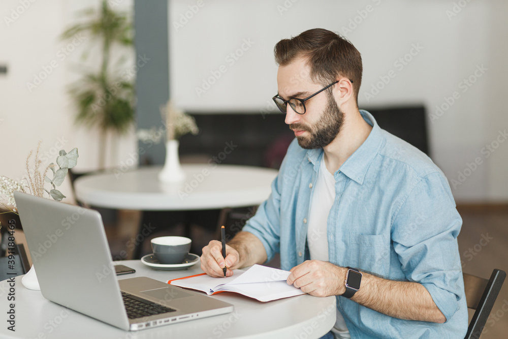 Serious young man in eyeglasses sitting alone at table in coffee shop cafe restaurant indoors working or studying on laptop pc computer writing in notebook. Freelance mobile office business concept. Serious young man in eyeglasses sitting alone at table in coffee shop cafe restaurant indoors working or studying on laptop pc computer writing in notebook. Freelance mobile office business concept.