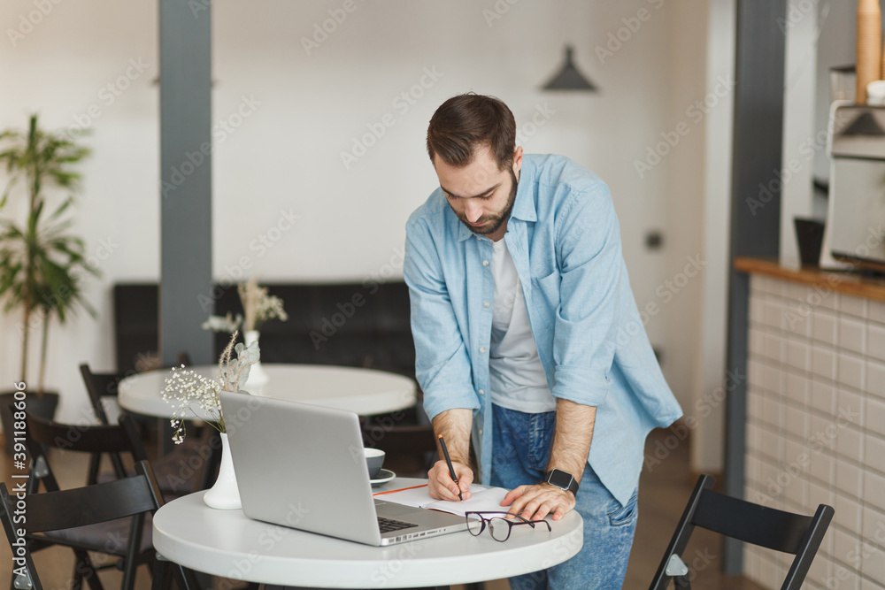 Handsome attractive young man standing alone near table in coffee shop cafe restaurant indoors working or studying on laptop pc computer writing in notebook. Freelance mobile office business concept.