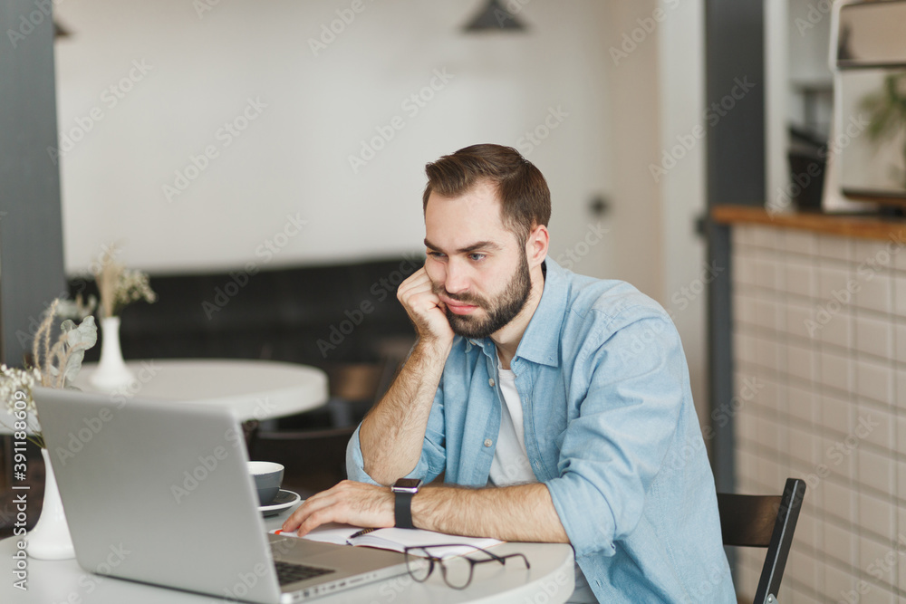 Dissatisfied boring young man sitting alone at table in coffee shop cafe restaurant indoor working or studying on laptop pc computer put hands on cheek. Freelance mobile office business concept. Dissatisfied boring young man sitting alone at table in coffee shop cafe restaurant indoor working or studying on laptop pc computer put hands on cheek. Freelance mobile office business concept.