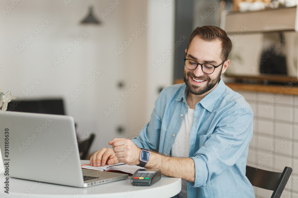 Smiling man in glasses sit at table in coffee shop cafe restaurant indoors working studying on laptop computer pay off with smart watch bank payment terminal. Freelance mobile office business concept. Smiling man in glasses sit at table in coffee shop cafe restaurant indoors working studying on laptop computer pay off with smart watch bank payment terminal. Freelance mobile office business concept.