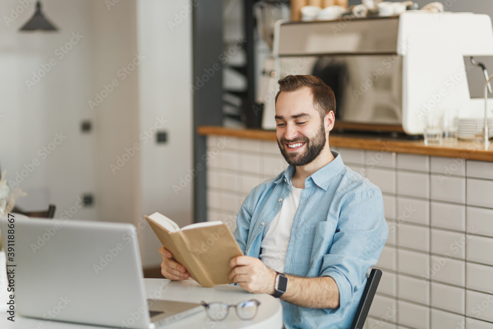 Smiling man sitting alone at table in coffee shop cafe working or studying on laptop computer reading book relaxing in restaurant during free time indoors. Freelance mobile office business concept.