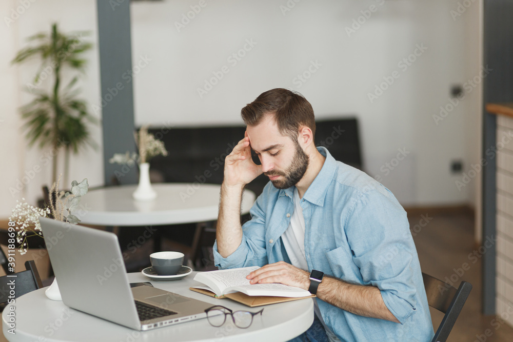 Handsome man sitting alone at table in coffee shop cafe working or studying on laptop computer reading book relaxing in restaurant during free time indoors. Freelance mobile office business concept. Handsome man sitting alone at table in coffee shop cafe working or studying on laptop computer reading book relaxing in restaurant during free time indoors. Freelance mobile office business concept.