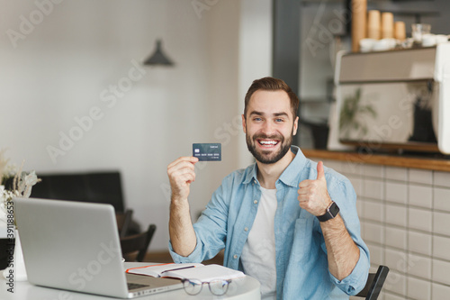 Smiling young man sit alone at table in coffee shop cafe restaurant indoor working or studying on laptop pc computer hold credit bank card showing thumb up. Freelance mobile office business concept.