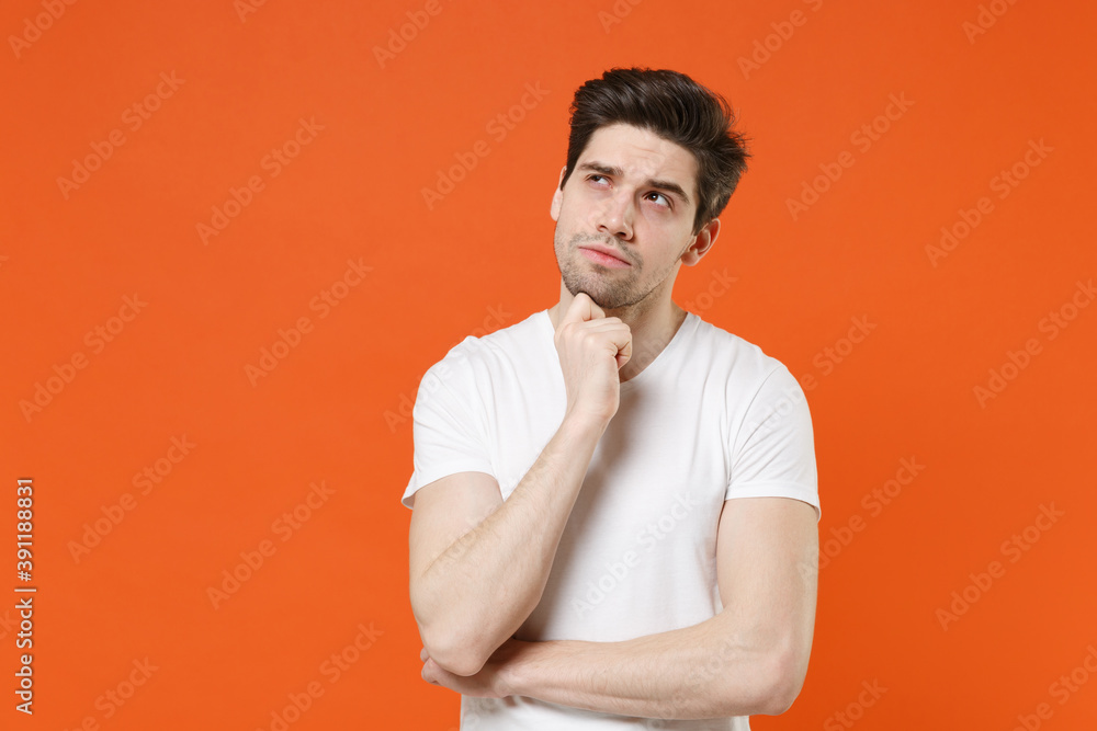 Pensive attractive handsome young man in basic casual white blank empty t-shirt standing put hand prop up on chin looking aside up isolated on bright orange colour wall background, studio portrait. Pensive attractive handsome young man in basic casual white blank empty t-shirt standing put hand prop up on chin looking aside up isolated on bright orange colour wall background, studio portrait.