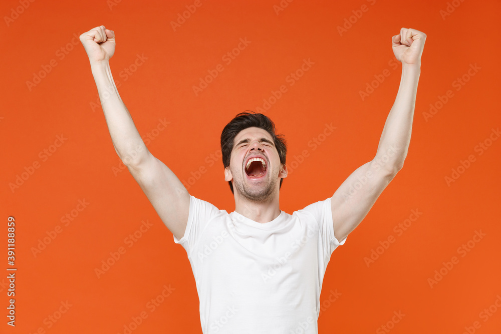 Fototapeta premium Overjoyed screaming young man in basic casual white blank empty t-shirt standing clenching fists doing winner gesture looking camera isolated on bright orange colour wall background, studio portrait.