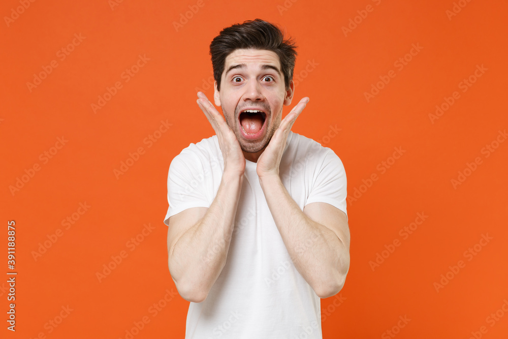 Shocked amazed surprised young man 20s in basic casual white t-shirt standing keeping mouth open put hands on cheeks looking camera isolated on bright orange colour wall background, studio portrait.