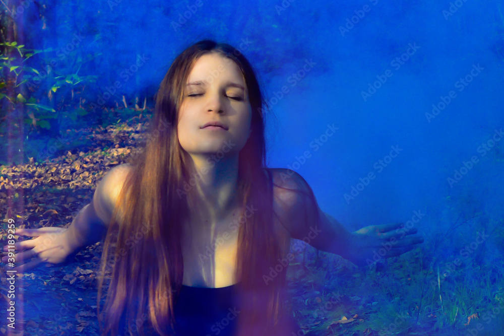 Red-haired woman sitting in a black long dress in a puff of blue smoke in the forest. Mysticism