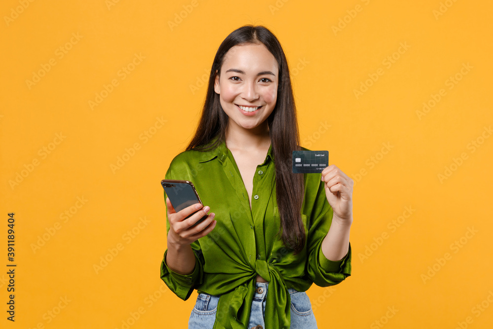 Smiling pretty young brunette asian woman wearing basic green shirt standing hold in hands using mobile cell phone hold credit bank card isolated on bright yellow colour background, studio portrait. Smiling pretty young brunette asian woman wearing basic green shirt standing hold in hands using mobile cell phone hold credit bank card isolated on bright yellow colour background, studio portrait.