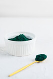Spirulina powder in a spoon and bowl on a white table.