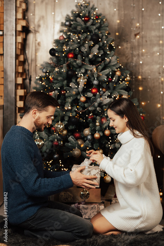 Charming couple in sweaters opening box with Christmas present. They celebrating holidays together in cozy home interior.