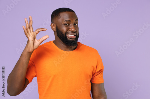 Confused puzzled young african american man 20s wearing basic casual orange t-shirt standing gesturing demonstrating size with workspace isolated on pastel violet colour background, studio portrait.
